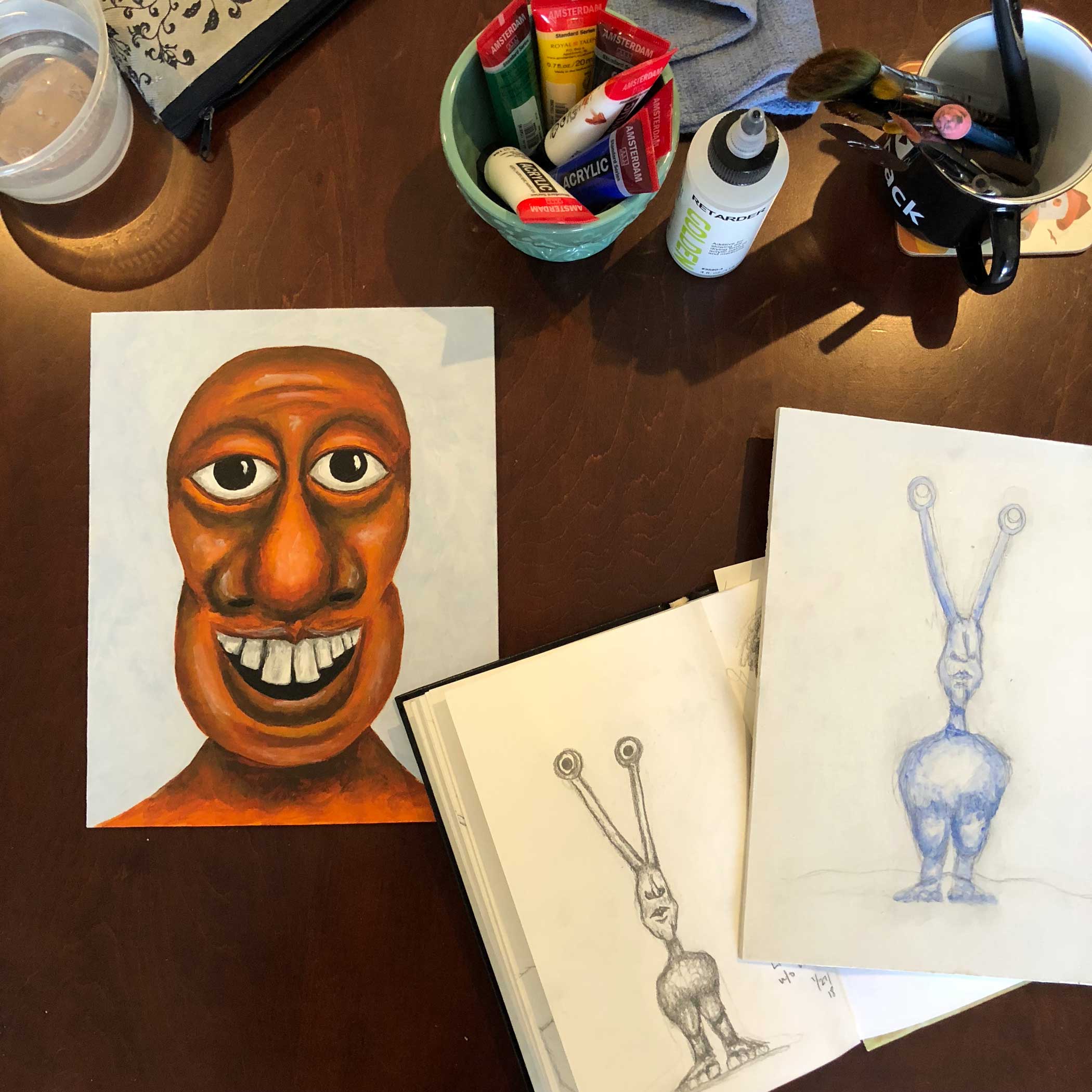 Finished orange monster portrait next to a painting in the beginning stages of a monster with weird antennae eyes and no arms