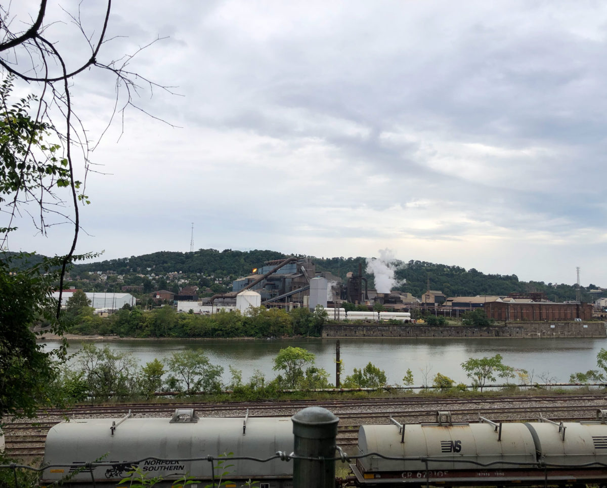 A view of the back of the steel factory, with train tracks visible and a flame coming from one of the stacks