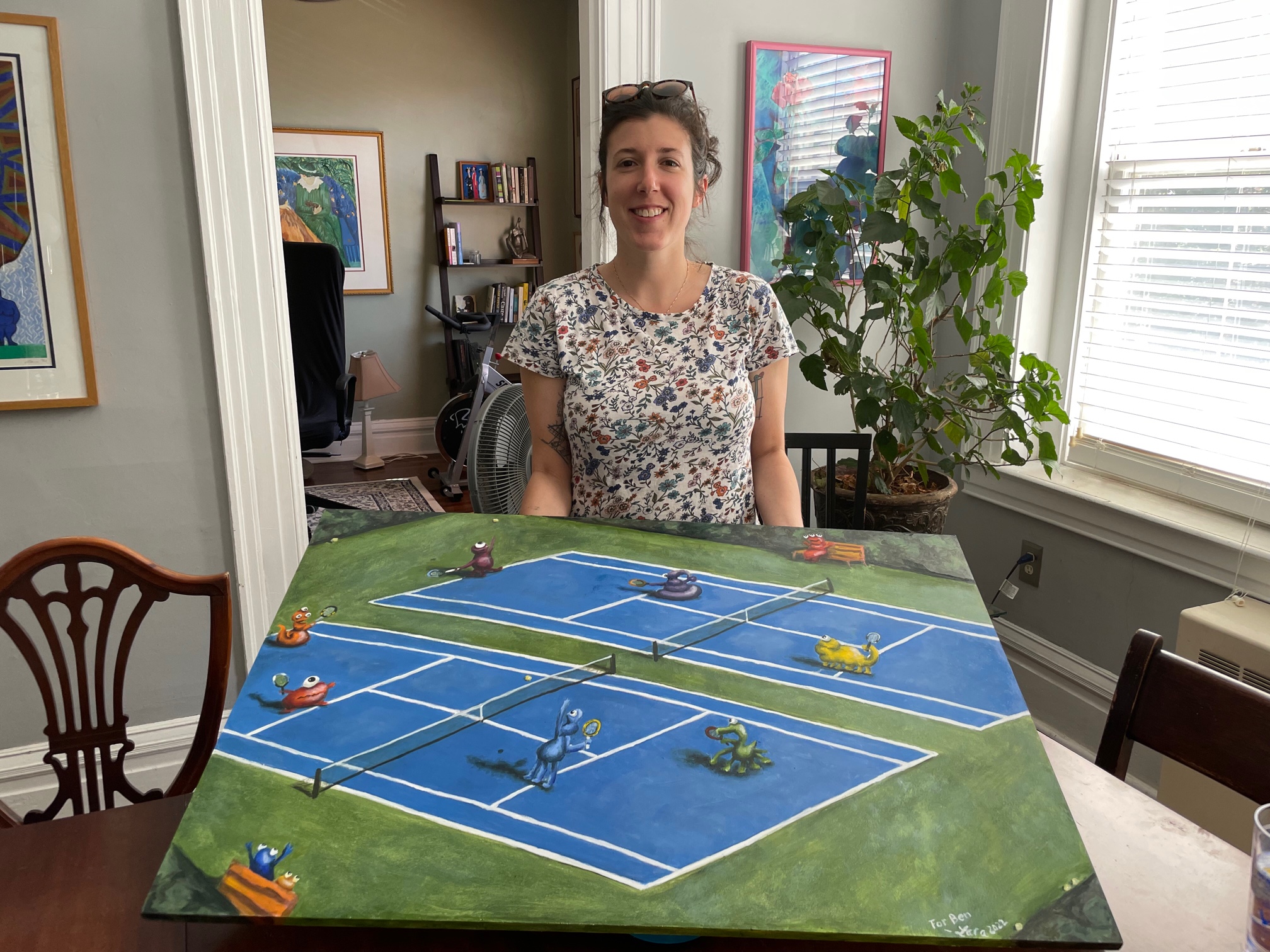 A white woman with brown hair and T Shirt with flowers on holding a large board with a painting of brightly colored small monsters playing tennis on two blue and green tennis courts. There are small benches with monsters viewing on the sides.